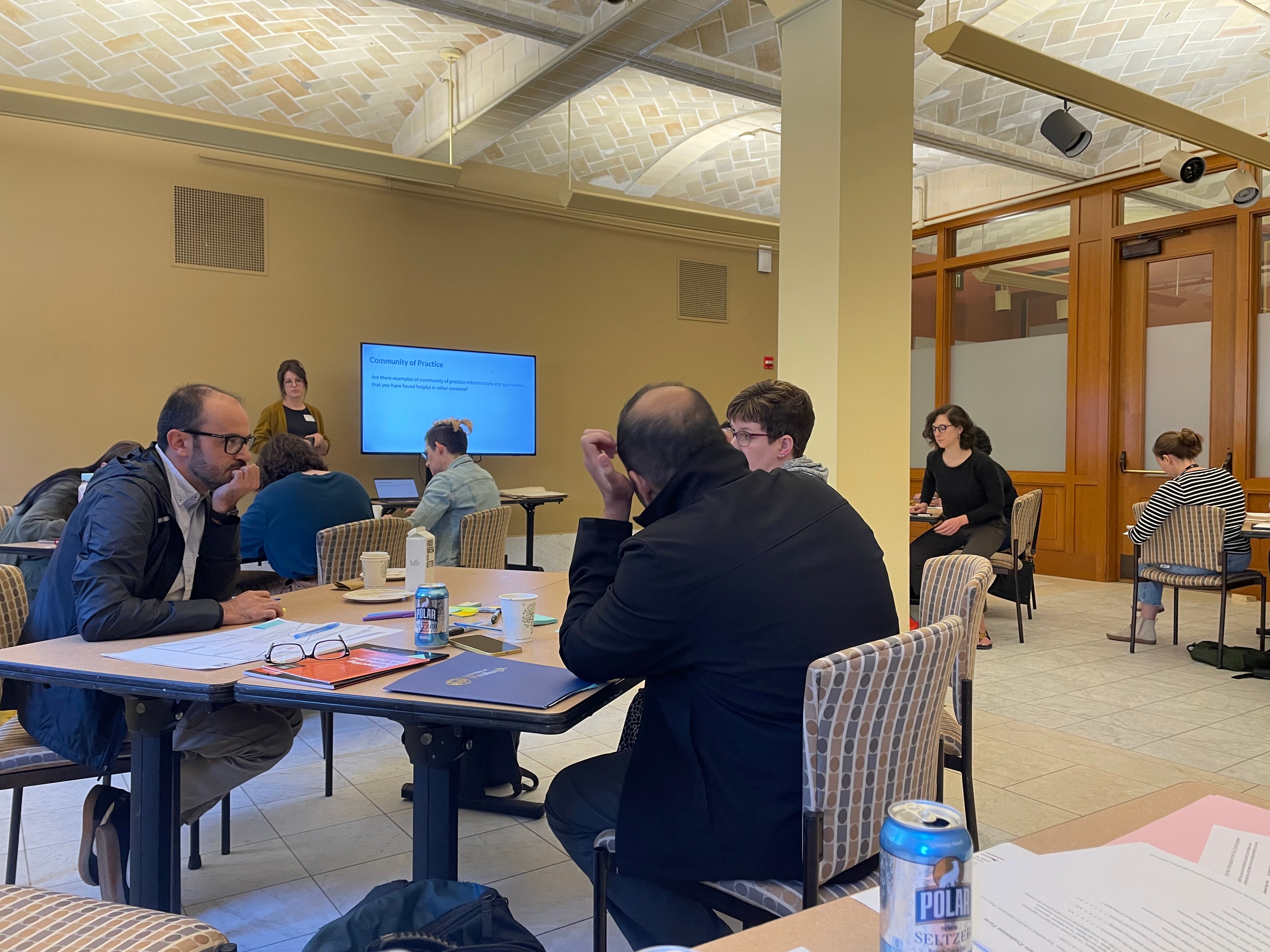 People sitting together at tables while someone presents a slideshow on a small screen