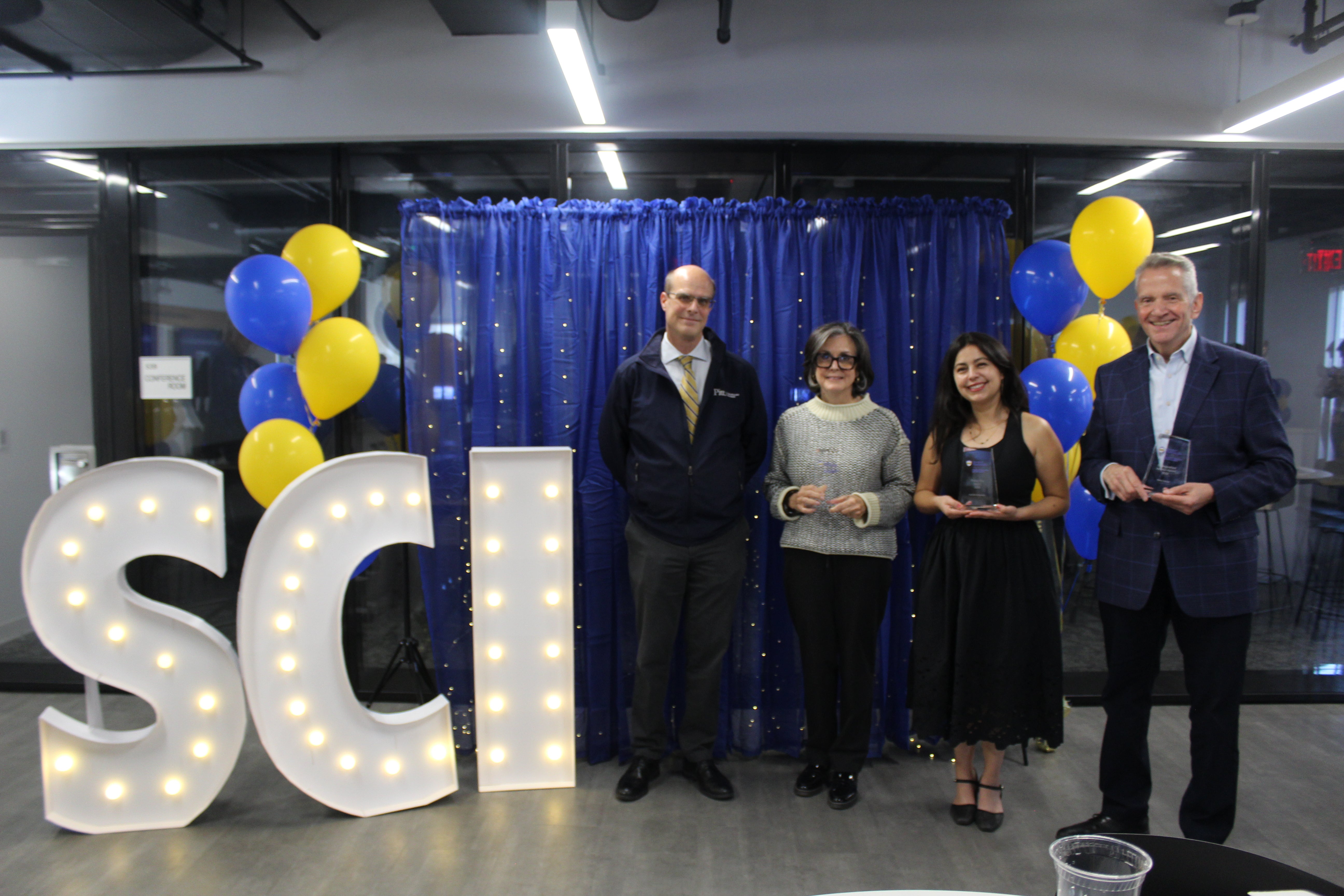 A photo of four people standing in front of a blue curtain and to the right of large light-up letters spelling out "SCI." Three of the four people are holding glass awards.
