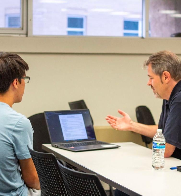 two men sitting at a table looking at a laptop