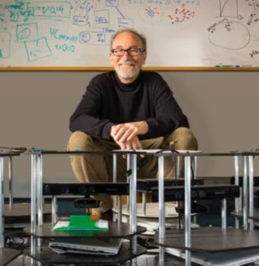 man sitting in front of white board wearing tan pants and a dark sweater
