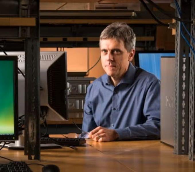 a man wearing a blue long sleeved shirt sitting at a desk next to a computer